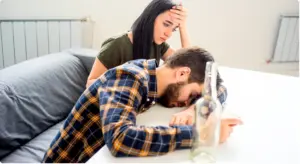 A distressed woman sitting on a sofa behind a man slumped over a table with an empty bottle, illustrating the personal impact of alcohol overconsumption.